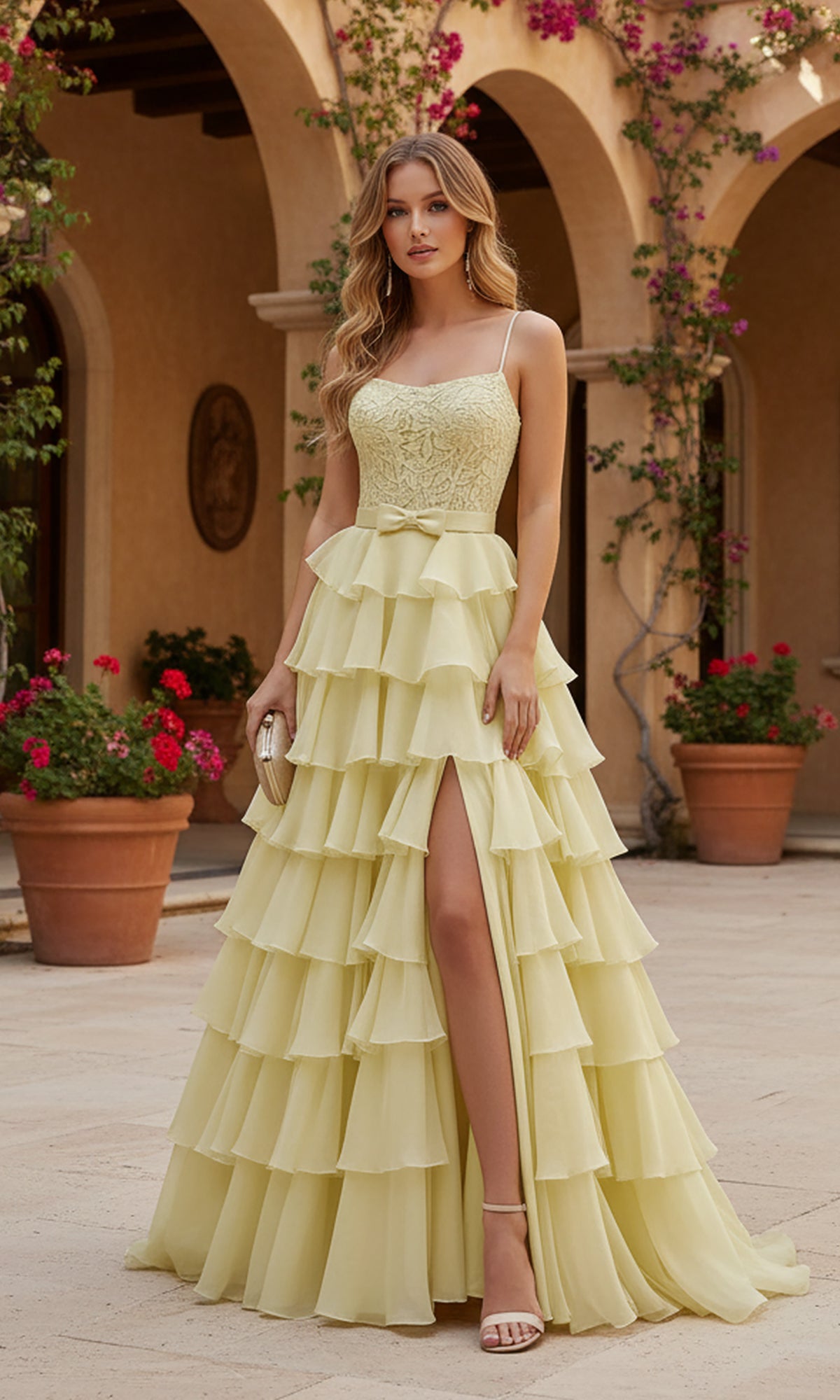 A woman stands outdoors wearing the PromGirl Exclusive 250202, a long light yellow tiered ruffle prom dress with a thigh-high slit, holding a small clutch amid potted plants and flowers.